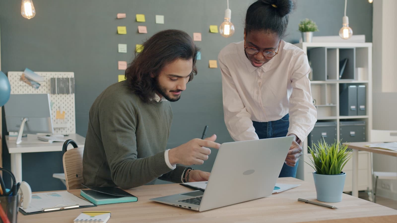 Two people using a laptop while reviewing work materials in an office.